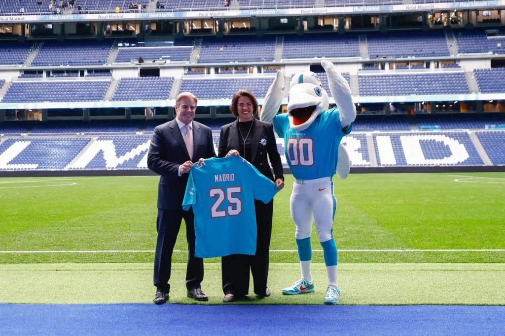 La NFL en Madrid trae de vuelta el alcohol al Bernabéu pese a la ley del deporte y dispara los alojamientos en la capital 1 Pri Shumate, directora de Marketing de los Miami Dolphins y Brett Gosper, director de la NFL en Europa y Asia-Pacífico, posan junto a la mascota de los Dolphins en el Santiago Bernabéu
