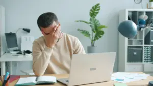 Joven abrumado en su escritorio con laptop, estrés de oficina.