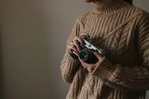 Una mujer con un suéter de punto sostiene una cámara vintage, mostrando un ambiente acogedor en el interior.